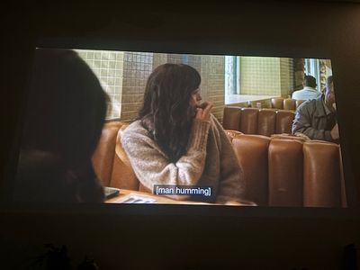 A woman across the table at a diner, turning to look over her shoulder. There are men solo at each booth behind her, and someone with a hood up sitting across from her.
