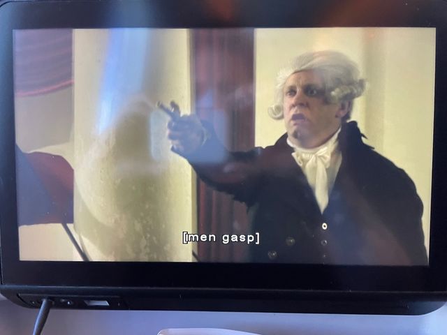 A man in period costume and a white wig seems upset, pointing a small period pistol off-screen
