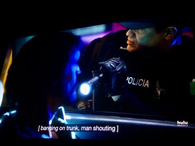 an officer in policia vest shines his flashlight into the car, lighting the driver, but he's looking back towards the trunk
