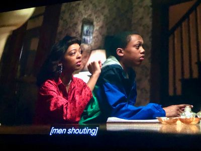 A teenager in red, and a child in a blue jacket sitting at a table, looking off right
