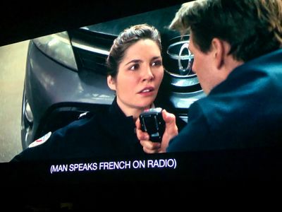 a police officer on the ground, with her back against the bumper of a car – a man in a suit kneels, holding her radio
