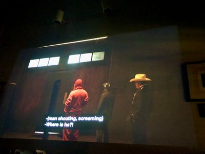 Interior, a sparse warehouse room with three men facing a door - one in a read sweatsuit and ski mask, one in black with a metal helmet, and one in a suit and cowboy hat
