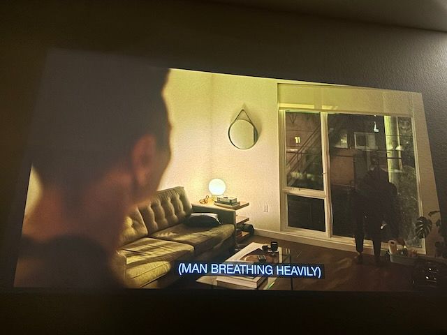 Over the shoulder of one man, an upstairs apartment with a sofa and glass coffee table. Someone is leaning over a bucket of cleaning supplies by the large window.
