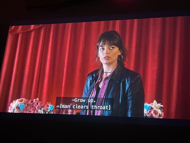 A teenager in a leather jacket glances to the side in front of red curtains and bouquets
