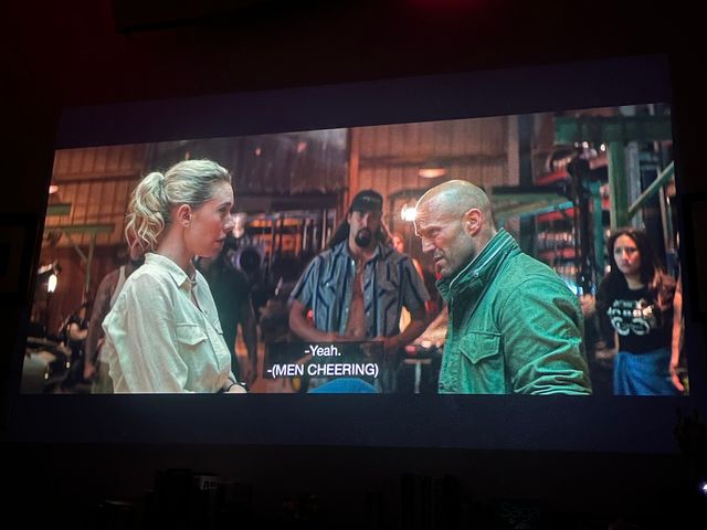 A blond woman and tough guy talking, he looks skeptical, as others look on
