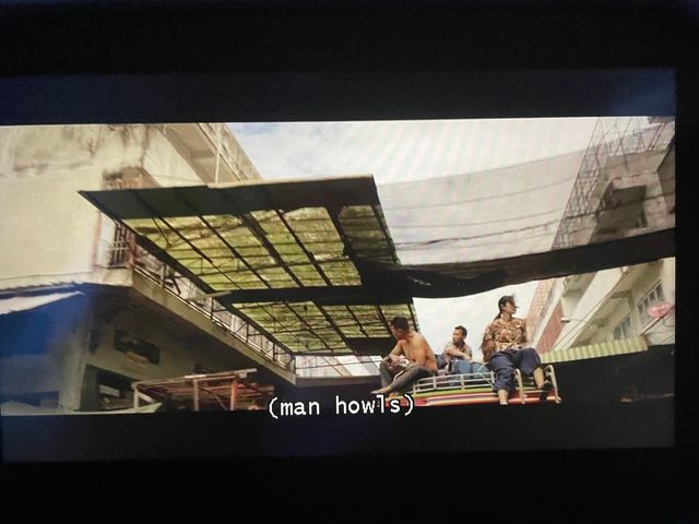 Three people sitting on the edge of a structure, with the tops of cement buildings behind them and some sort of awning
