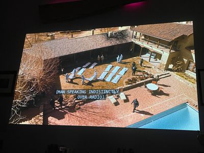 From far above, the courtyard and pool of a large house, with white body bags laid out in rows - police officers and people in hazmat stand around or move bodies
