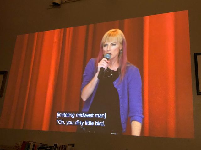 Maria Bamford with a microphone in front of a red curtain, she's wearing a blue cardigan and has blond hair dyed pink from the ears down
