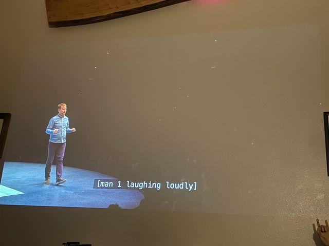 A comedian on a thrust stage walking to one side as we can see glimpses - past him, the audience in darkness
