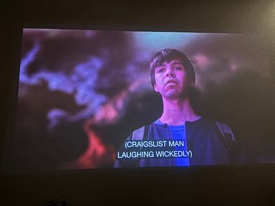 Young man in bowl cut and backpack above us looking down, framed by storm clouds or a volcano, reds and blues and billowing black
