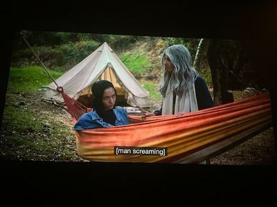 Young woman sitting up in an orange hammock glaring at an older guru-like woman standing beside her. There's a white tent behind them, and nature all around
