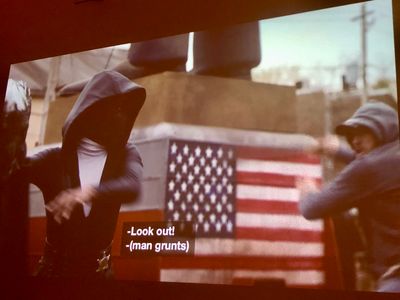 Exterior, two figures fighting in front of an american flag - a woman in black-and-white hood and mask with a sheriff badge on her hip, and a man in a baseball cap and hoodie
