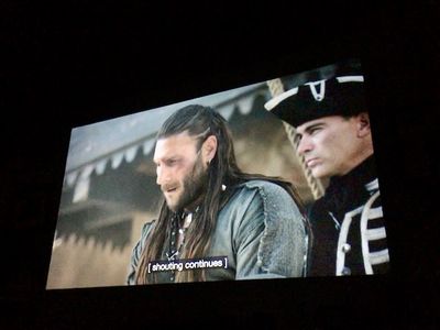 A man with long hair pulled back wearing a rough pirate tunic, talking to a man in black and white uniform with a naval hat
