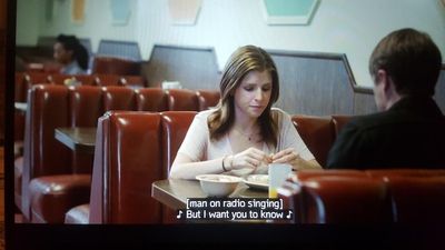 Young woman with long hair in a diner booth handling at her food, not looking at the man across -- his back to us
