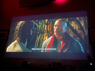 The Rock in a black and red scarf talking to a woman with braids, we only see the back of her head
