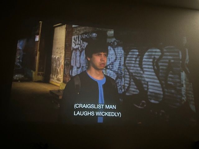 Young man with bowl cut and backpack looking disgusted. He's on a dark street at night, with graffiti lit behind him
