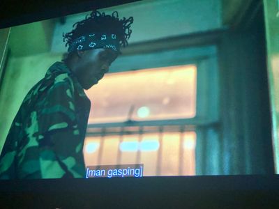 A young Black man in a cement hallway, backlit by a window, wearing a camouflage jacket and green bandana
