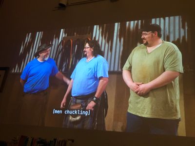 Three smiling men standing side by side in t-shirts against a metal shop wall - left man in a black cowboy hat is congratulating center man in a tartan kilt, as right man in a cap looks on
