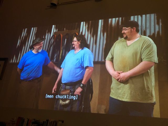 Three smiling men standing side by side in t-shirts against a metal shop wall - left man in a black cowboy hat is congratulating center man in a tartan kilt, as right man in a cap looks on
