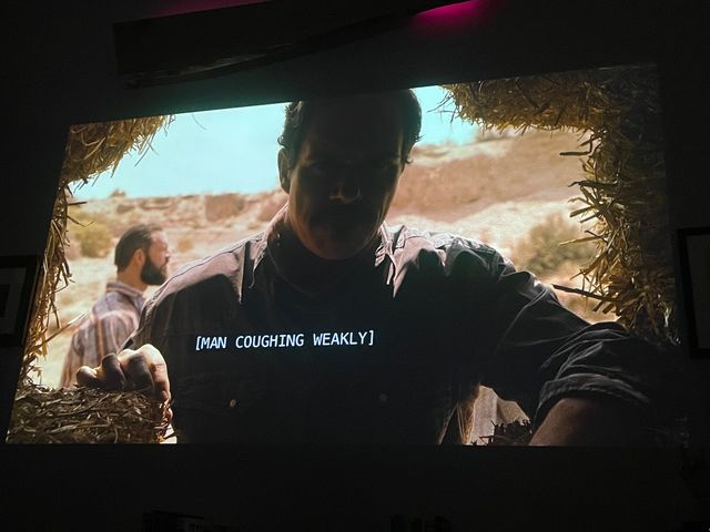 A man framed by hay bales and backlit by a desert sun looks at the camera, face in shadow - another man behind him
