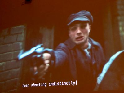 external against brick, a distraught young man in a period cap and jacket points a pistol past the camera
