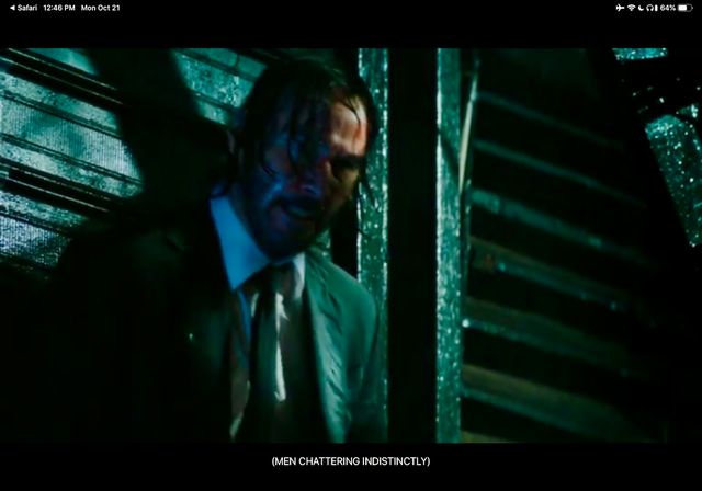 Keanu drenched in a suit and tie with his back to a metal wall and covered in dramatic shadows
